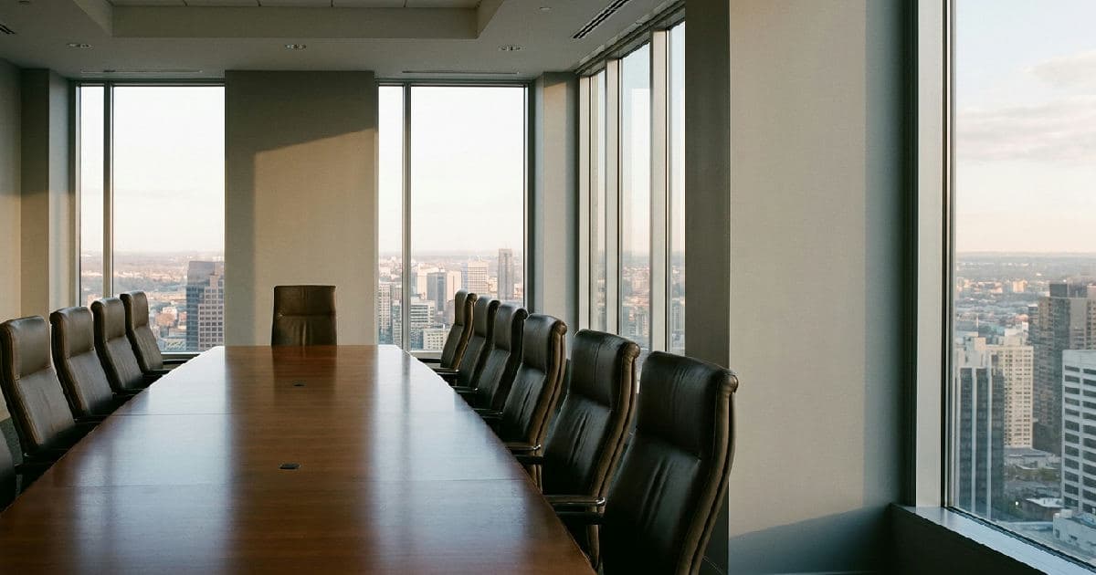 Vacant boardroom chair at head of table with city view beyond windows.