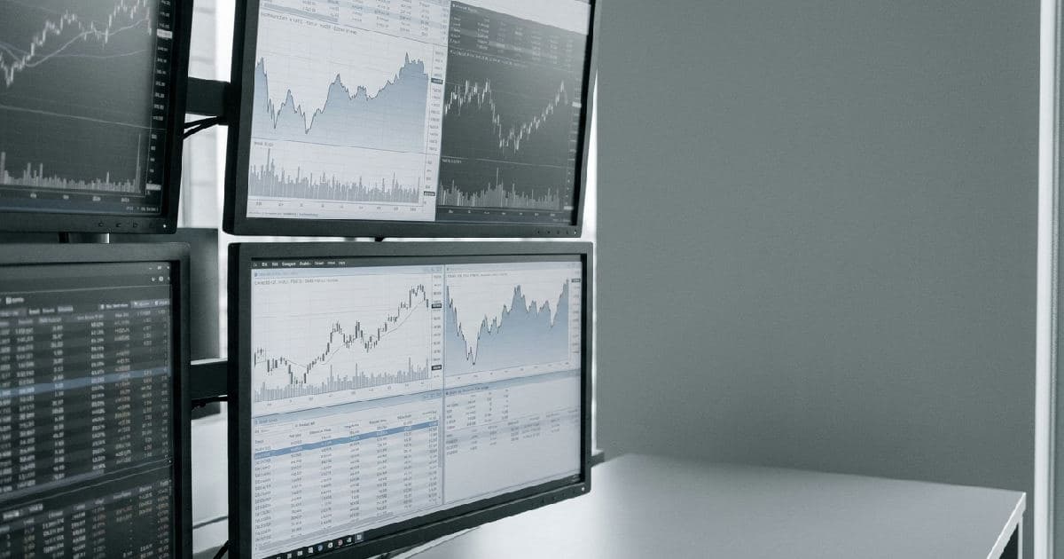 Computer monitors displaying financial market charts and data in a desaturated, professional setting.