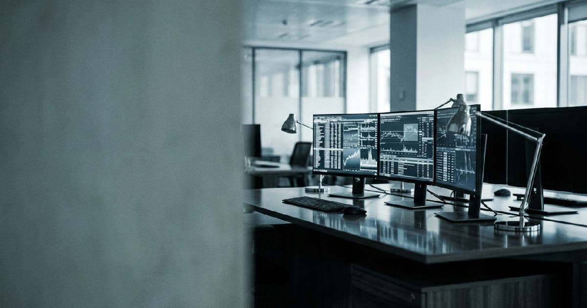 Trading desk with multiple monitors showing financial data visualisations and market charts in muted tones.