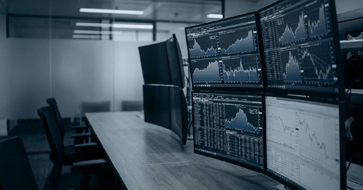 Multiple computer monitors displaying financial market data and analytical charts in a trading environment.