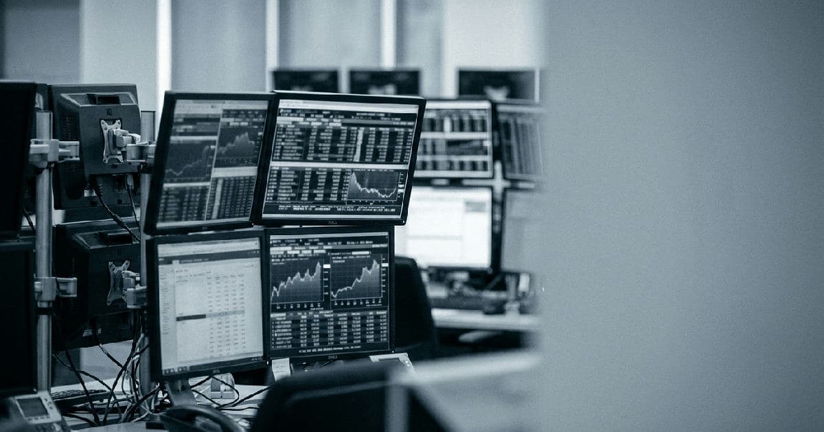 Multiple monitors showing financial data and market charts in muted tones, trading floor setting.
