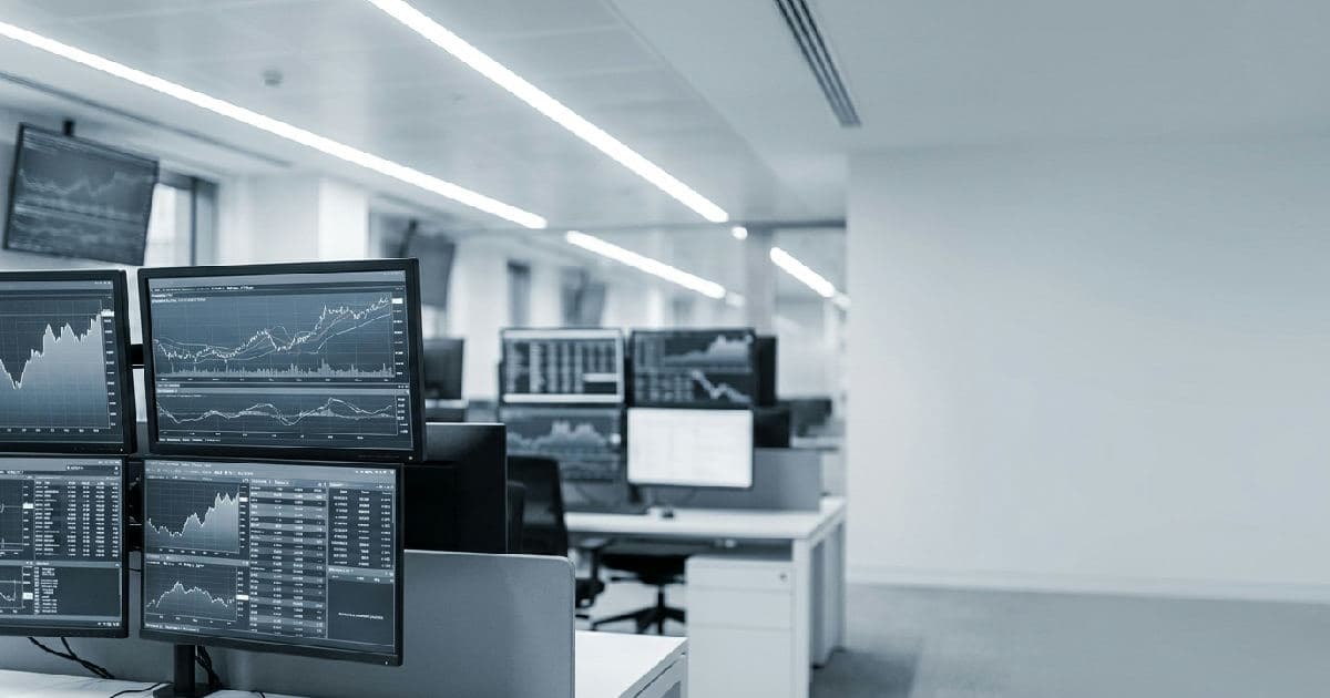 Trading floor with multiple screens showing financial data visualisations in muted tones, no people visible.